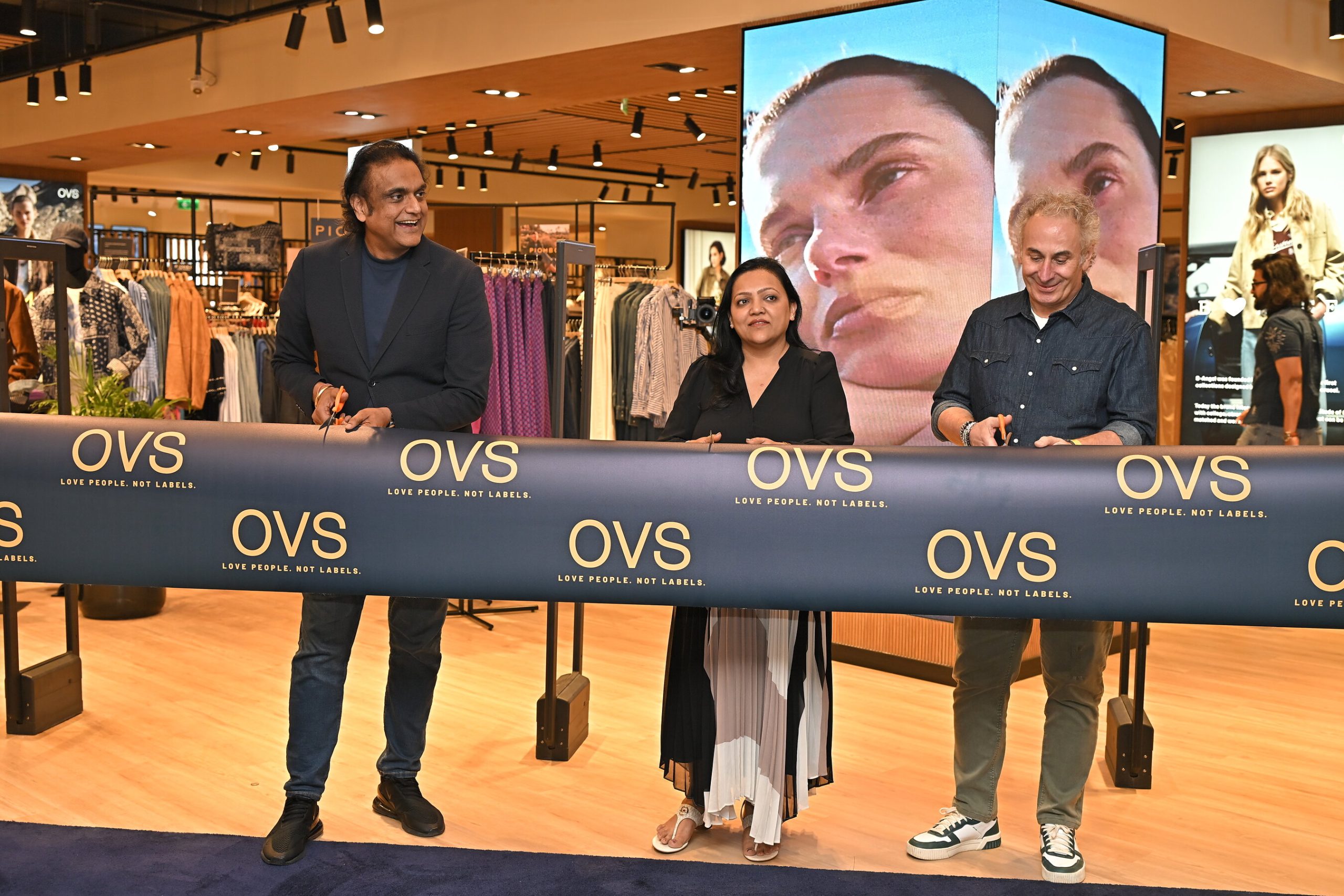 Three people cut a navy OVS ribbon at the opening of the OVS store in Sky City Mall, Mumbai, with clothing racks and a video wall behind them.