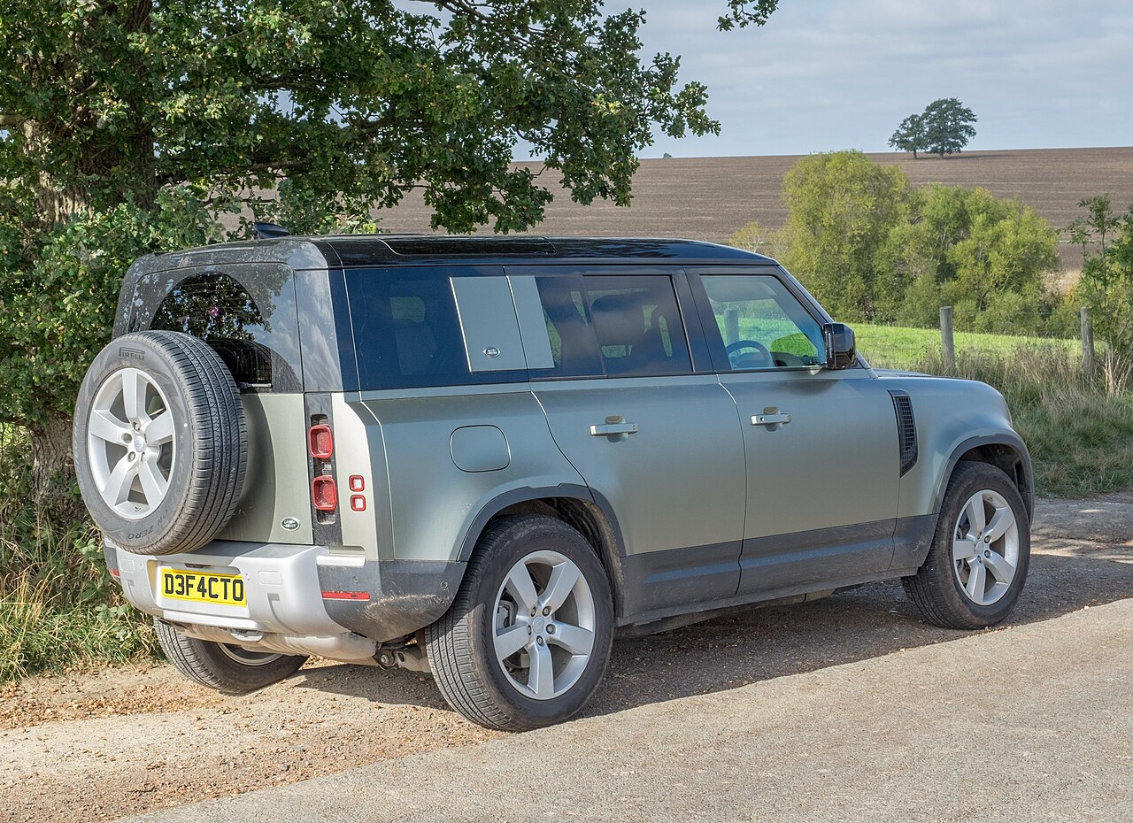 Rear three-quarter view of a gray-green Land Rover Defender 110 with a spare wheel on the tailgate, parked on a country road